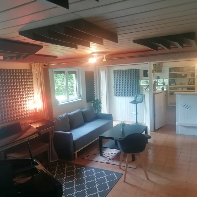 Vue large du salon acoustique : piano à queue, canapé gris, table basse et cuisine ouverte, plafond équipé de pièges à basses